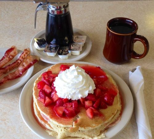 A plate of pancakes topped with strawberries and whipped cream, served with bacon, syrup, and a cup of coffee.