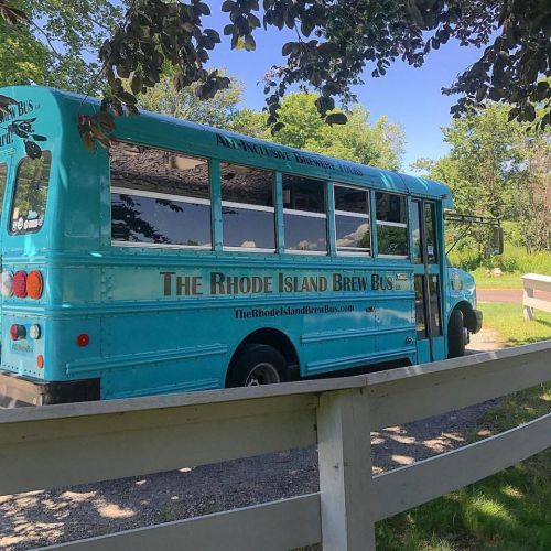 A turquoise bus with "The Rhode Island Brew Bus" written on the side, parked near a white fence in a green area.