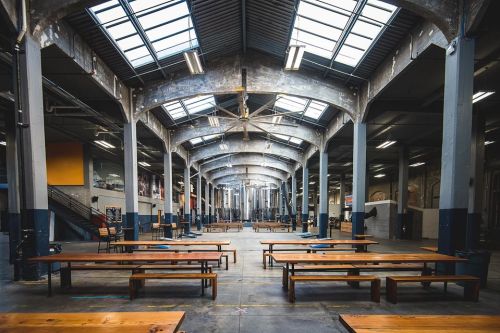 Spacious industrial hall with high ceilings, large windows, and long wooden tables arranged in rows.
