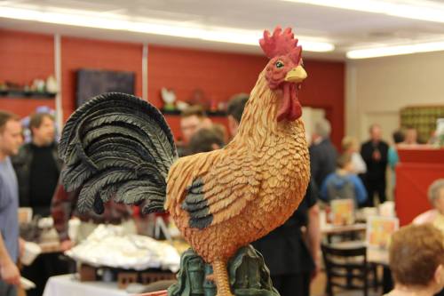 A detailed rooster statue in the foreground, with a busy restaurant scene in the background.