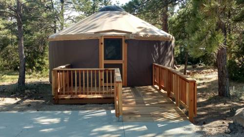 A wooden yurt with a tan canvas top, featuring a ramp and surrounded by trees in a natural setting.