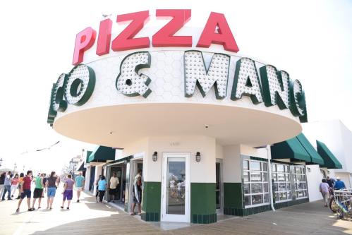 A vibrant pizza restaurant with a large sign, bustling with people on a sunny day.
