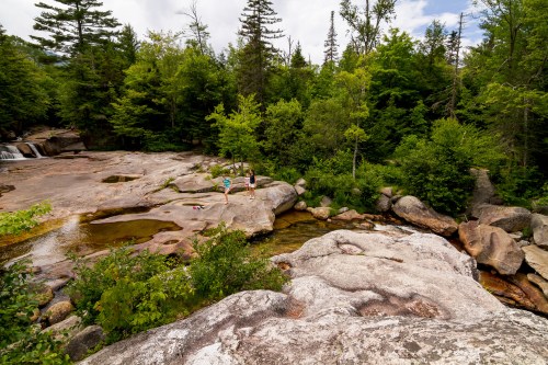 Two people stand on rocky terrain by a stream, surrounded by lush green trees and a cloudy sky.