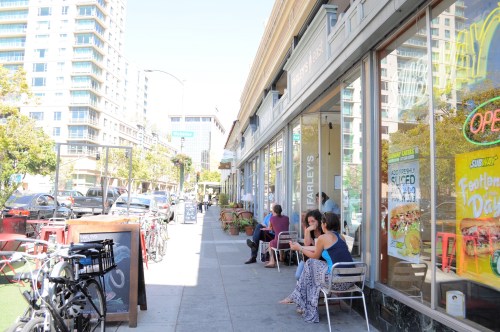 A sunny street scene with people sitting outside a café, bicycles parked, and buildings in the background.