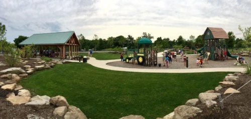 A panoramic view of a playground with children playing, surrounded by green grass and trees under a cloudy sky.