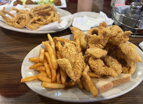 A plate of crispy fried chicken tenders on toast, served with golden French fries, with onion rings in the background.