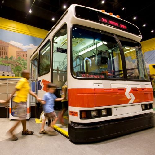 Children play near a colorful bus exhibit in a museum, with a vibrant mural in the background.