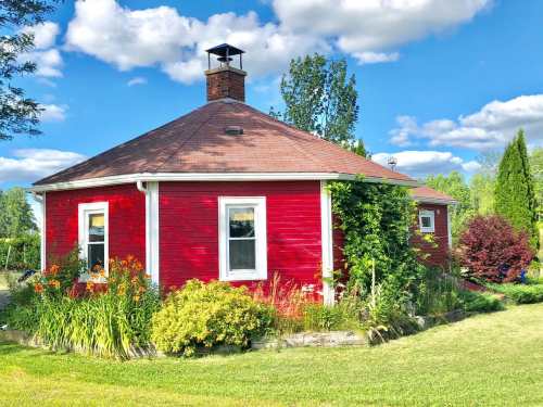 A round red house surrounded by lush greenery and colorful flowers under a bright blue sky with fluffy clouds.