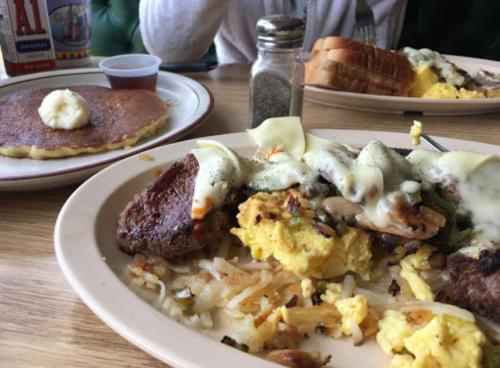 A plate of breakfast featuring scrambled eggs, hash browns, steak, and a side of pancakes with butter.