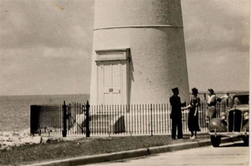 Two women converse near a lighthouse, while a man in uniform stands nearby, with a vintage car parked on the road.