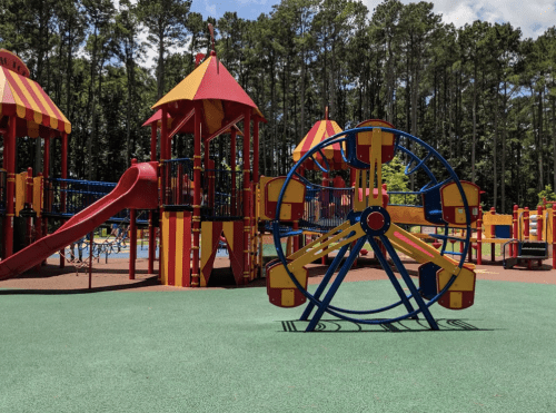 Colorful playground with a slide, climbing structures, and a small Ferris wheel, surrounded by trees and blue sky.