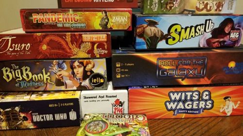 A collection of board games stacked on a table, including titles like Pandemic, Smash Up, and Wits & Wagers.