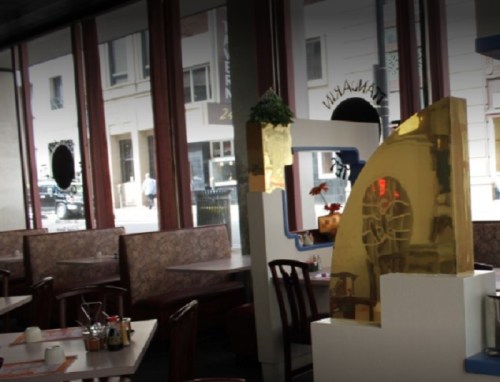 Interior of a restaurant featuring wooden chairs, tables, and a decorative golden structure. Natural light from windows.