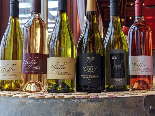 A display of six bottles of Pfeiffer wine, including varieties like Musky, Pink, Pinot Gris, and Chardonnay, on a wooden table.