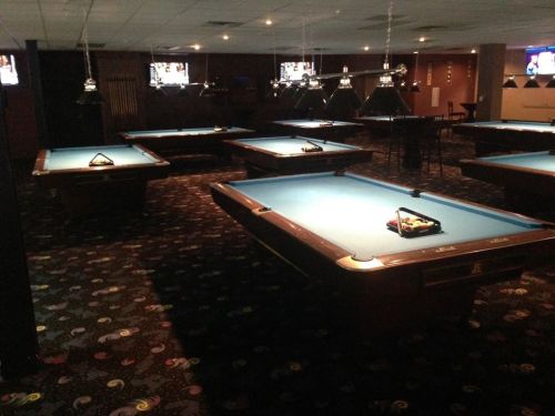 A dimly lit pool hall featuring several blue felt pool tables and overhead lights.