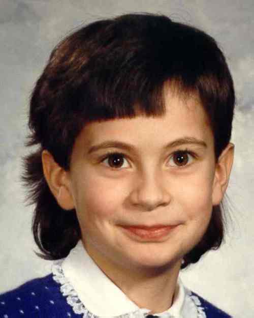 A young girl with dark hair and a mullet hairstyle smiles for a school photo, wearing a blue sweater and white collar.