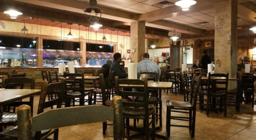 A casual restaurant interior with wooden tables and chairs, featuring a few diners and a view of the kitchen area.