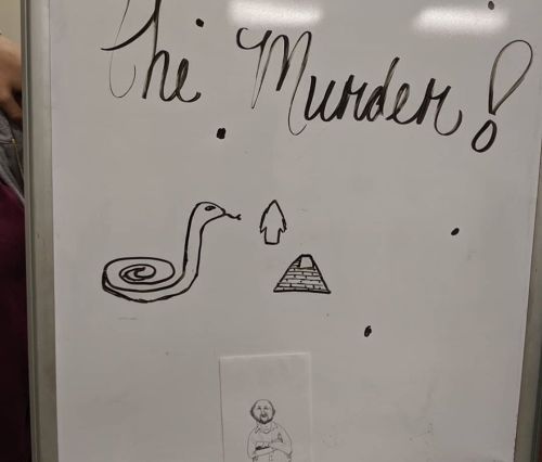 A whiteboard with the text "The Murder!" and drawings of a snake, an arrow, a pyramid, and a small figure.