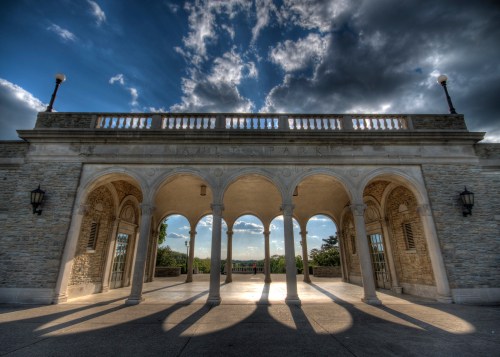 A stone pavilion with arched columns under a cloudy sky, casting long shadows on the ground.