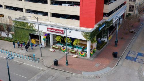 Aerial view of a colorful bar with outdoor seating, located near a parking garage and street with pedestrians.
