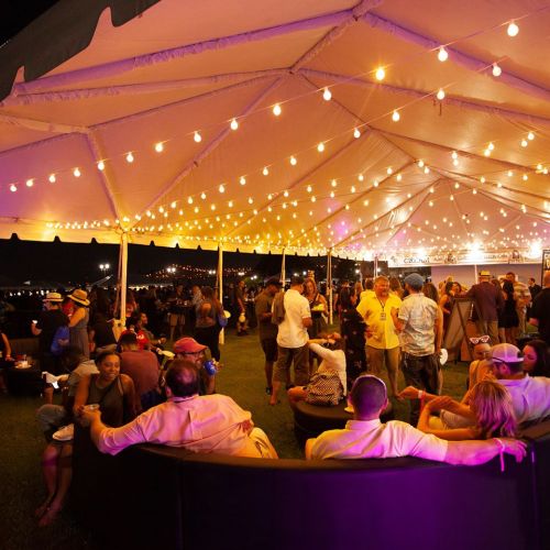 A lively outdoor event under a tent with string lights, featuring guests socializing and enjoying the atmosphere.