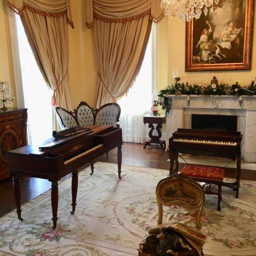 A vintage room featuring two pianos, elegant drapes, a chandelier, and a decorative rug.