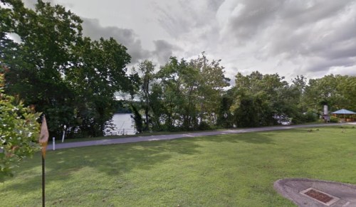 A grassy area with trees and a river in the background, under a cloudy sky. A path runs alongside the water.