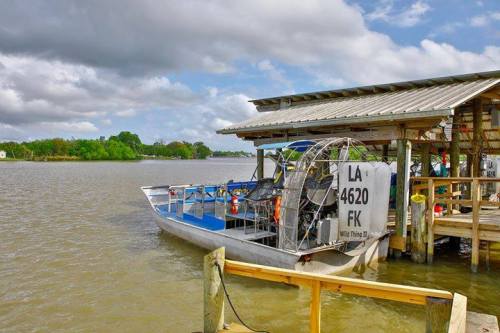 An airboat docked at a wooden pier on a calm river, surrounded by greenery and cloudy skies.