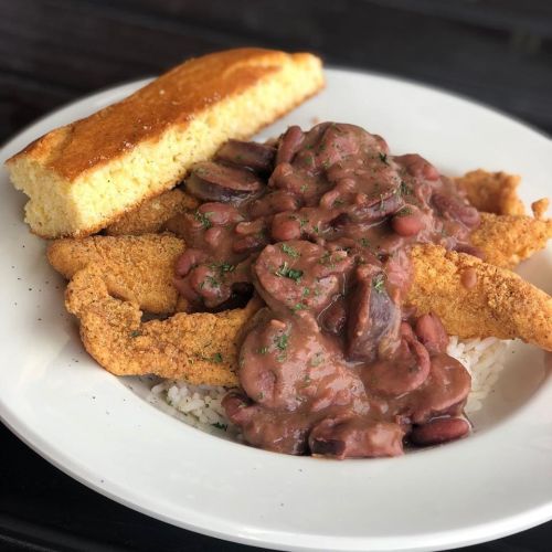 A plate of fried fish fillets topped with red beans and rice, served with a slice of cornbread.