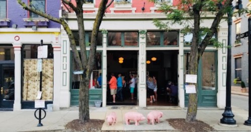 Three pigs stand outside a colorful storefront where people are gathered inside. Trees frame the scene.