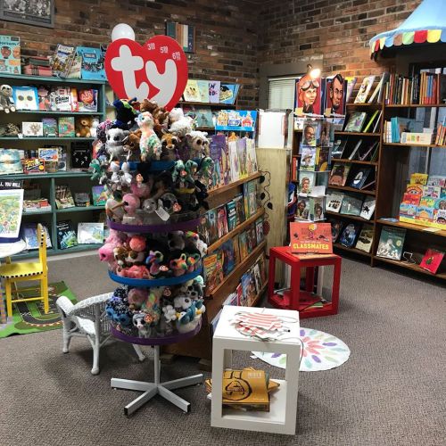 A cozy bookstore featuring a display of plush toys, colorful bookshelves, and a reading area with chairs and rugs.