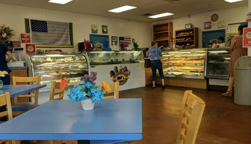 A cozy bakery interior with display cases of pastries, a blue table, and customers browsing treats. American flag on the wall.