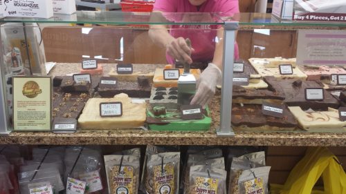A person in a pink shirt serves fudge from a display case filled with various chocolate treats and snacks.