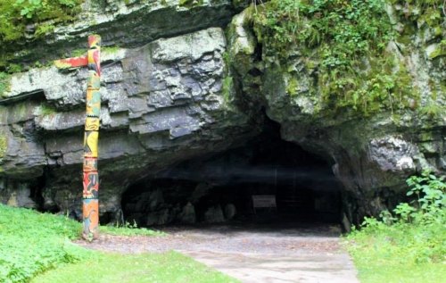 A cave entrance surrounded by lush greenery, featuring a colorful totem pole on the left.