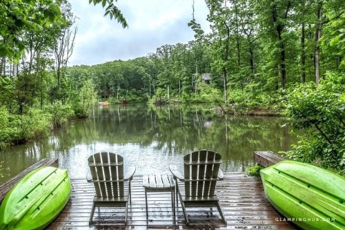 Two green kayaks and three wooden chairs overlook a serene, misty lake surrounded by lush greenery.