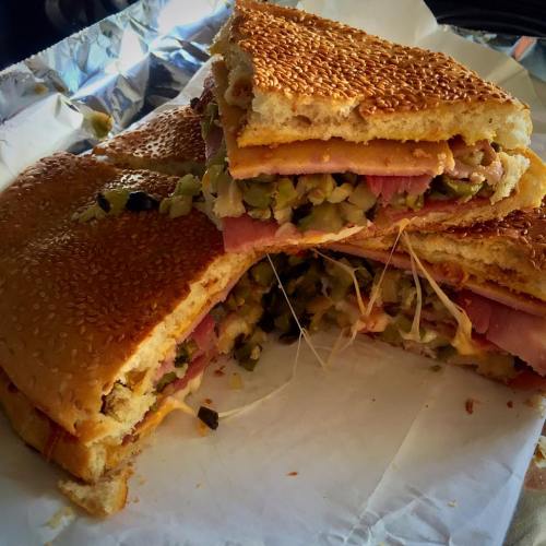 A large sandwich with sesame seed bread, layers of ham, cheese, and pickles, cut into sections and served on foil.