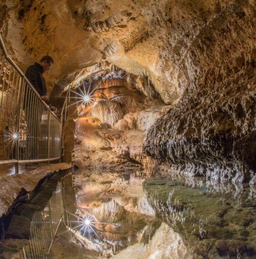 A person stands by a railing in a cave, reflecting on still water surrounded by illuminated rock formations.