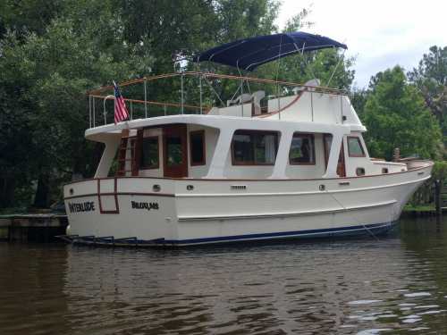 A white boat named "Interlude" docked on calm water, surrounded by lush greenery and trees.