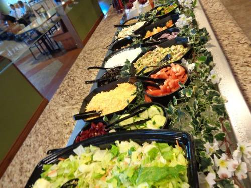 A buffet-style salad bar with various toppings, including greens, cheese, tomatoes, and dressings, on a granite counter.