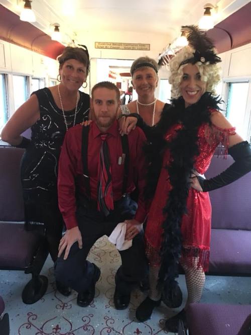 Four people dressed in 1920s-style outfits pose together inside a vintage train car, smiling and enjoying the moment.
