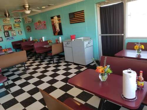 A retro diner interior with checkered floors, maroon tables, and flower centerpieces, featuring a trash bin and wall decor.