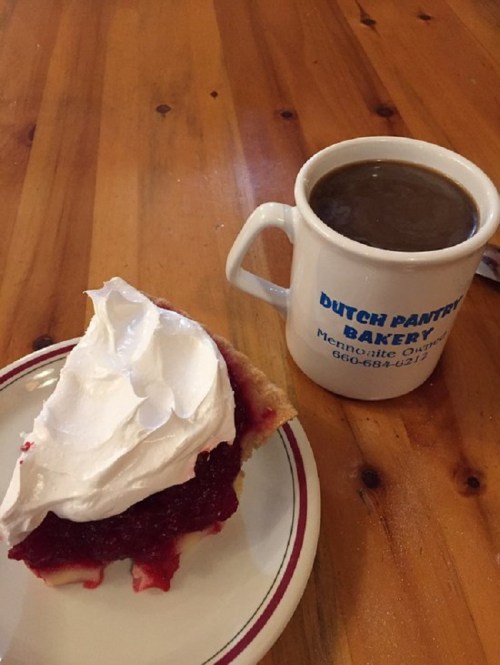 A slice of cherry pie topped with whipped cream next to a cup of coffee on a wooden table.