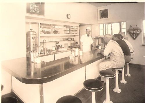 A vintage diner scene with a counter, a smiling waiter, and two customers seated on stools, enjoying drinks.