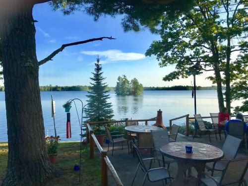 A serene lakeside view with trees, a patio area, and calm water reflecting the sky.