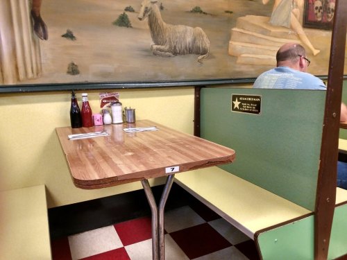 A cozy diner table with condiments, silverware, and a mural of a llama in the background. A person sits nearby.