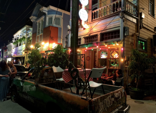 A vibrant street scene at night featuring a colorful restaurant, a vintage truck, and people enjoying the atmosphere.