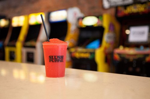 A vibrant red slush drink in a cup with "Slush Puppie" logo, set against a backdrop of arcade machines.