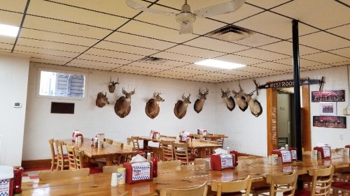 A rustic dining area with wooden tables, chairs, and mounted deer heads on the walls, featuring a restroom sign.