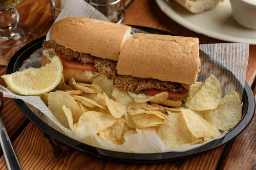 A crispy chicken sandwich on a bun with tomato, served with a side of potato chips and a lemon wedge.
