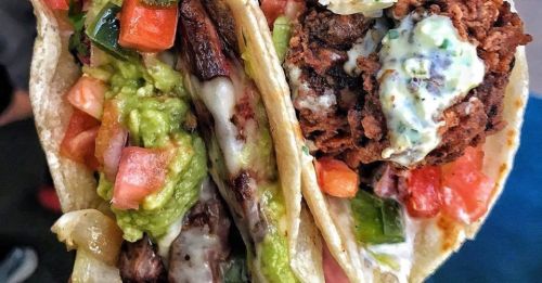 Close-up of two tacos filled with grilled meat, guacamole, fresh tomatoes, and creamy sauce, served in soft tortillas.